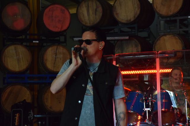 Singer in sunglasses at the mic, barrels stacked behind the stage